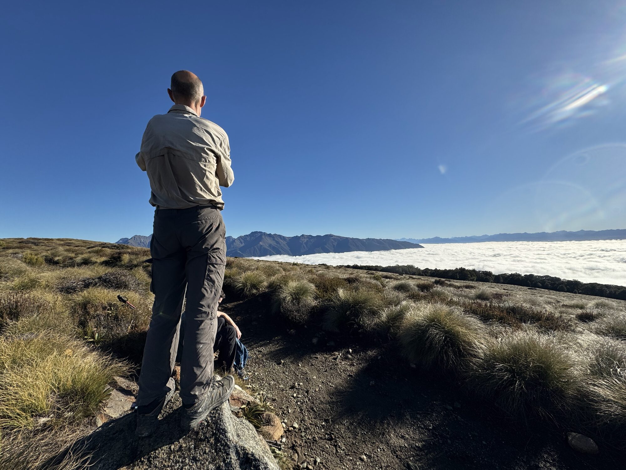 Vic, seen from behind, standing on a rock above a sea of cloud with mountain ridges on the horizon; someone sits on the ground behind him.