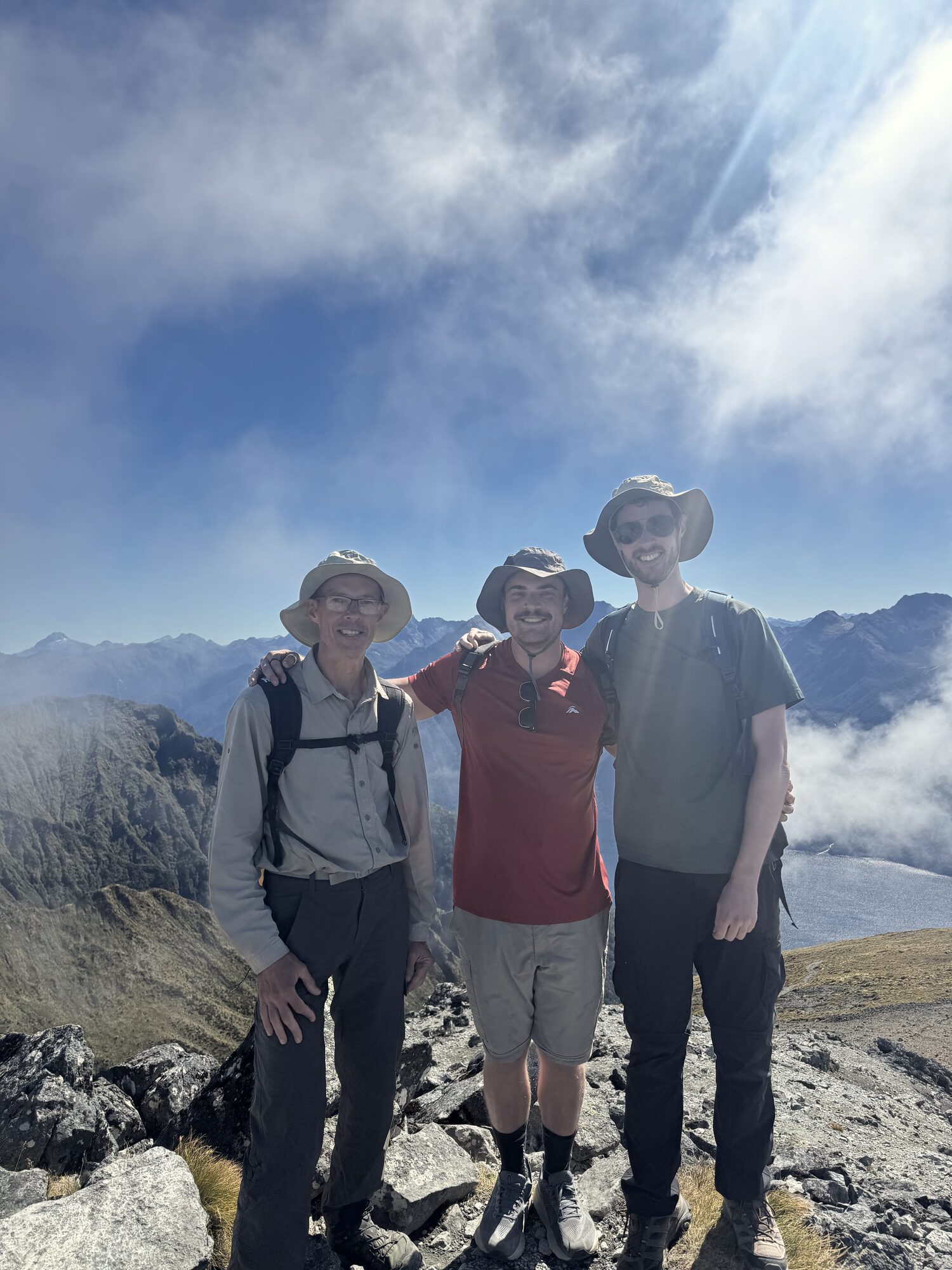 Vic in a sun hat with two younger companions on a rocky summit, cloud and mountains behind them.