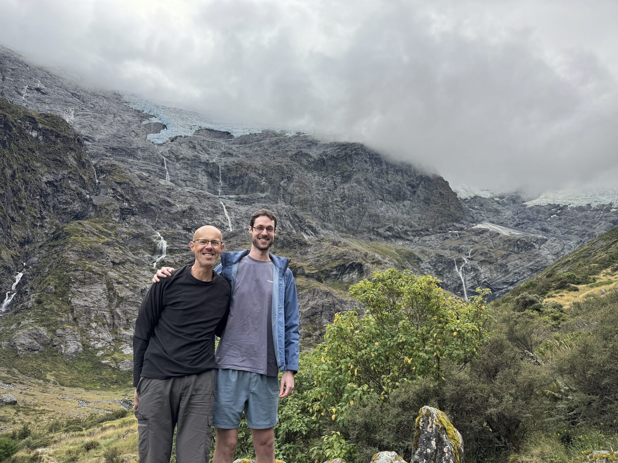 Vic and a younger man standing arm in arm below a mountain wall with waterfalls, cloud over the peaks.